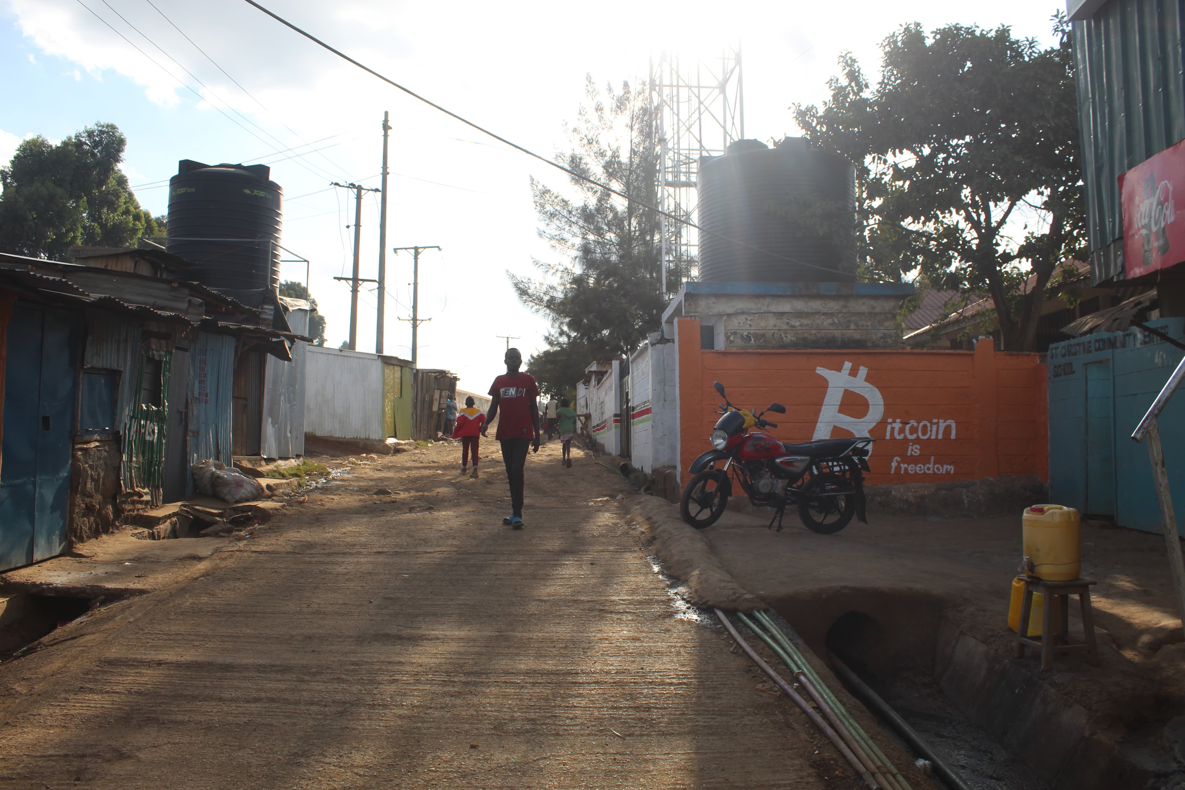 A street in Soweto West, Kibera — the Bitcoin is freedom mural visible in the background alongside a boda-boda motorcycle and residents walking